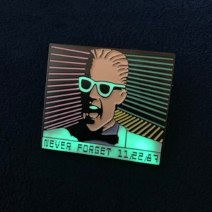 Puede incluir: Un pin de esmalte negro y verde con un personaje de dibujos animados que lleva gafas de sol y una pajarita. El personaje está delante de un fondo a rayas verde neón, rosa y amarillo. El texto "NEVER FORGET 11/22/87" está impreso debajo del personaje.
