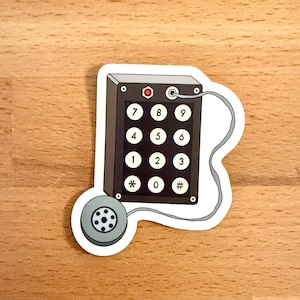 Könnte beinhalten: Ein Aufkleber mit einer Retro-Telefon-Tastatur und einem Hörer. Die Tastatur ist schwarz mit weißen Zifferntasten und einem roten Licht. Der Hörer ist grau mit einem kreisförmigen Lautsprecher. Der Aufkleber hat einen weißen Rand.