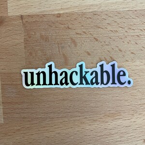 Könnte beinhalten: Ein holografischer Aufkleber mit dem Wort "unhackable" in fetter, schwarzer Schrift. Der Aufkleber hat einen perlmuttartigen Glanz, der einen Regenbogen von Farben reflektiert. Der Aufkleber ist rechteckig mit abgerundeten Kanten.