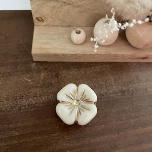 Peut inclure: Bouton décoratif en forme de fleur à quatre pétales, blanc, avec des accents dorés. Le bouton est posé sur une surface en bois foncé, avec une étagère en bois et de petites sphères décoratives en arrière-plan.