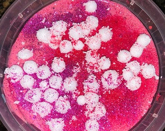 May include: A container of pink slime with red and pink glitter and white foam balls. The slime is in a clear plastic container. The glitter is spread throughout the slime, and the foam balls are scattered on top.