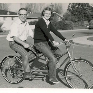 May include: Black and white photograph of two people riding a tandem bicycle on a paved road. The bicycle has a classic design with fenders and a high handlebar. The people are wearing casual clothing, and the background shows houses and trees.