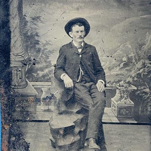 May include: Antique black and white portrait of a man seated outdoors. He wears a dark suit, hat, and a decorative chain. The background features a painted landscape with architectural elements, creating a vintage aesthetic.