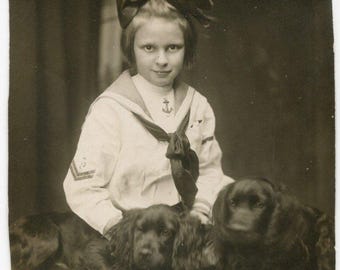 Young girl wearing a sailor-style outfit and hair bow, with two Spaniel dogs, c 1910. Vintage original real photo studio print.