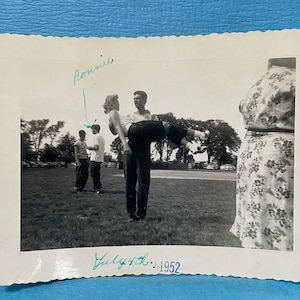May include: Black and white photograph of a man holding a woman in his arms, with other people in the background. The photo is dated July 4th, 1952, and has the name "Ronnie" written on it.
