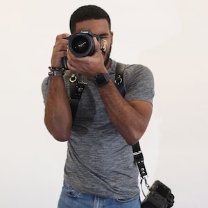 Puede incluir: Una persona con una camiseta gris y pantalones vaqueros azules sostiene una c&aacute;mara negra con un objetivo. La c&aacute;mara est&aacute; unida a una correa de cuero negra.