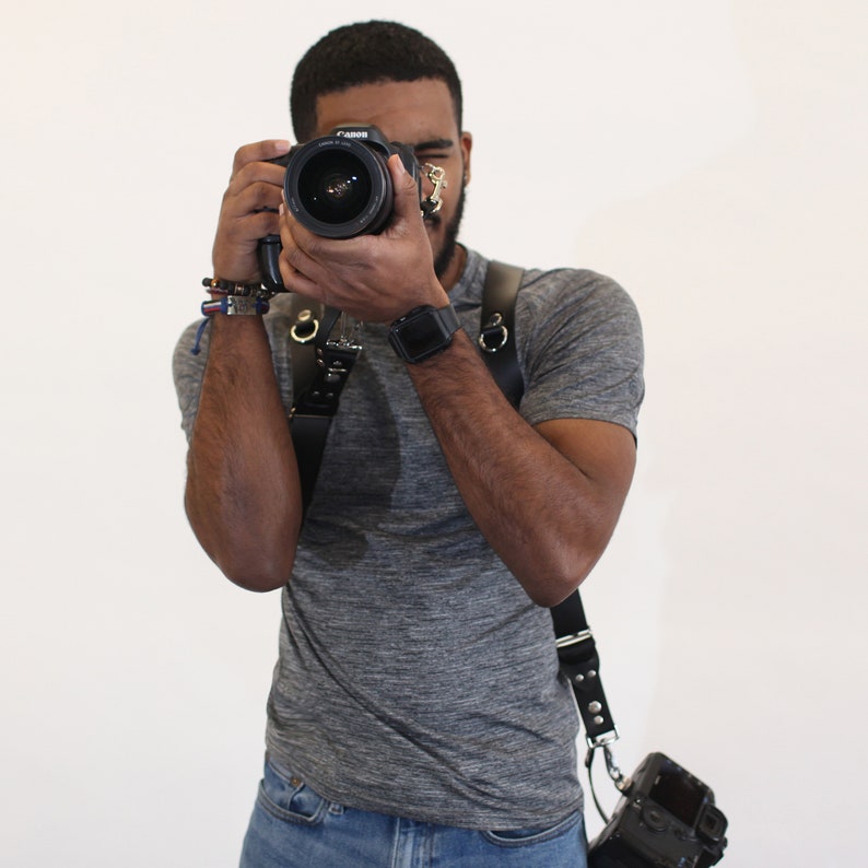 Puede incluir: Una persona con una camiseta gris y pantalones vaqueros azules sostiene una c&aacute;mara negra con un objetivo. La c&aacute;mara est&aacute; unida a una correa de cuero negra.