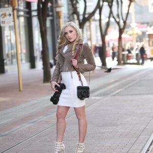 Puede incluir: Una mujer con una chaqueta de gamuza marr&oacute;n y un vestido blanco, sosteniendo una c&aacute;mara con una correa de cuero marr&oacute;n. Est&aacute; de pie en una calle de la ciudad.