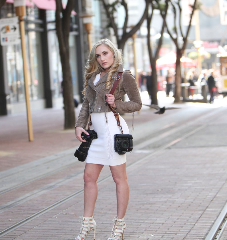 Puede incluir: Una mujer con una chaqueta de gamuza marr&oacute;n y un vestido blanco, sosteniendo una c&aacute;mara con una correa de cuero marr&oacute;n. Est&aacute; de pie en una calle de la ciudad.