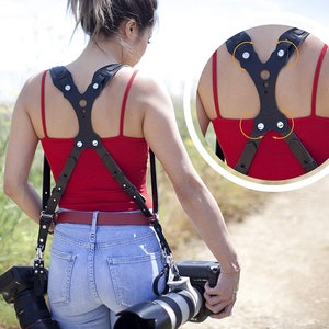 May include: A black leather camera strap with adjustable straps and a padded back panel. The strap is designed to distribute weight evenly across the shoulders and back.