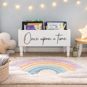 May include: A white wooden bookshelf with the words "Once upon a time" in black script holds several children's books. A rainbow-shaped rug and a star-shaped light add to the whimsical decor. A teddy bear sits in a basket.