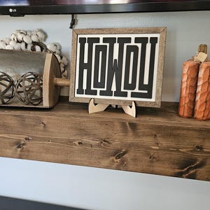May include: A wooden shelf displays decorative items. A framed sign with the word "HOWDY" in black, a carved pumpkin, a clock, and a metal scoop with decorative spheres are visible. The shelf is made of dark wood.