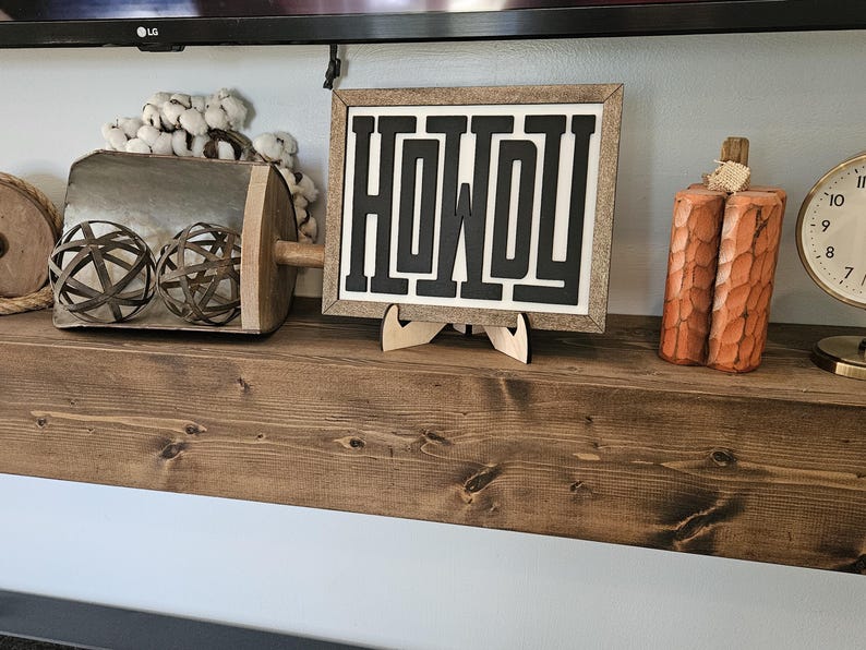 May include: A wooden shelf displays decorative items. A framed sign with the word "HOWDY" in black, a carved pumpkin, a clock, and a metal scoop with decorative spheres are visible. The shelf is made of dark wood.