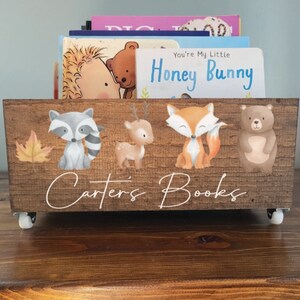 May include: A wooden book bin with woodland animal illustrations, including a raccoon, deer, fox, and bear. The bin is brown with the text "Carter's Books" and sits on small wheels. Books are visible inside.
