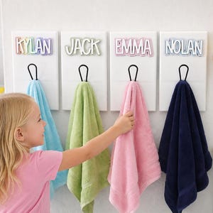 May include: Four white rectangular wall-mounted towel hooks, each featuring a personalized name cutout in a different color: Kylan in rainbow, Jack in green, Emma in pink, and Nolan in blue. Each hook holds a matching colored towel.