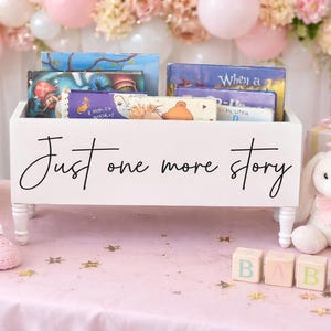 May include: A white wooden book bin with the text "Just one more story" holds several children's books. The bin sits on a pink surface with baby-themed decorations, including a stuffed bunny, baby shoes, and blocks that spell "BABY".