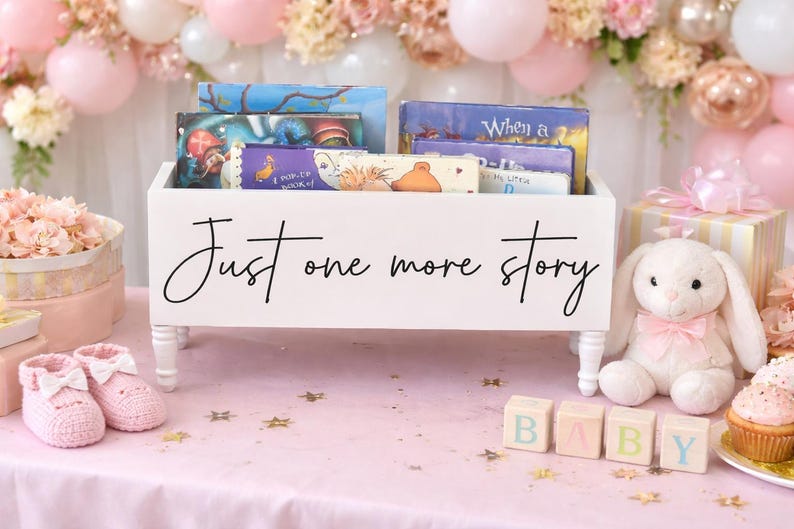 May include: A white wooden book bin with the text "Just one more story" holds several children's books. The bin sits on a pink surface with baby-themed decorations, including a stuffed bunny, baby shoes, and blocks that spell "BABY".