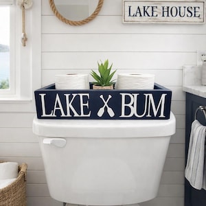 May include: A navy blue wooden box with the words "LAKE X BUM" in white lettering, placed on a white toilet. The box holds two rolls of white toilet paper and a small green plant. A "Lake House" sign is visible.