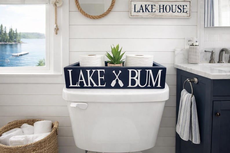 May include: A navy blue wooden box with the words "LAKE X BUM" in white lettering, placed on a white toilet. The box holds two rolls of white toilet paper and a small green plant. A "Lake House" sign is visible.