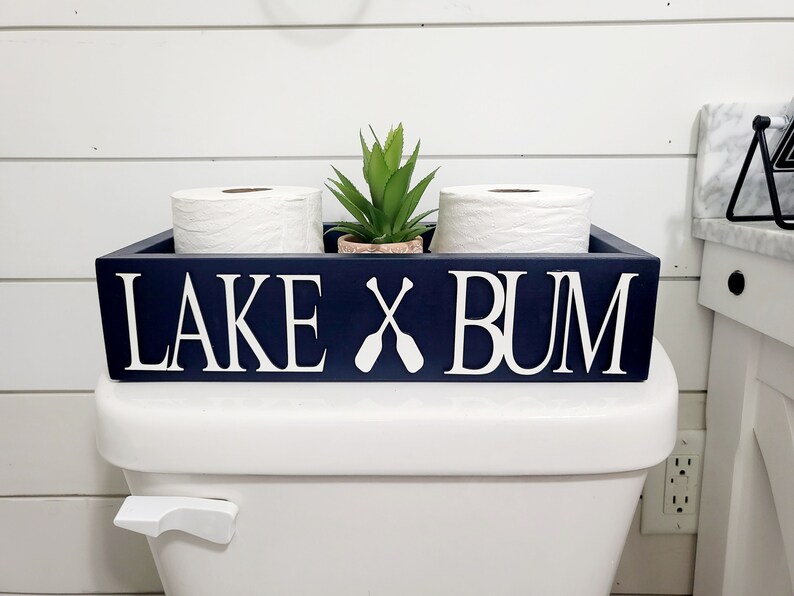 May include: A navy blue wooden toilet paper holder with the words "LAKE X BUM" in white. The holder contains two rolls of white toilet paper and a small potted plant. The design includes crossed oars.