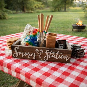 May include: A rectangular dark wood box with the white text S'mores Station and a graphic of two crossed roasting sticks. The box contains a clear glass jar holding four wooden roasting sticks, a bag of marshmallows, and chocolate bars.