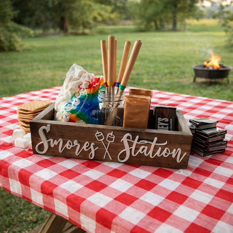 May include: A rectangular dark wood box with the white text S'mores Station and a graphic of two crossed roasting sticks. The box contains a clear glass jar holding four wooden roasting sticks, a bag of marshmallows, and chocolate bars.