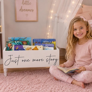 May include: A white wooden book bin with four short legs, featuring the black cursive text "Just one more story" on the front. The bin is filled with various colorful children's books, providing a storage solution for a bedroom or playroom.