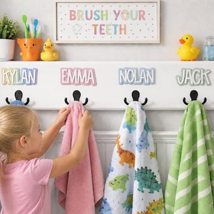 May include: A white wall-mounted shelf with hooks, personalized with names. The shelf holds towels, a toothbrush holder, and a sign that says "Brush Your Teeth." A young girl is hanging a pink towel.