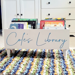 May include: A white wooden bookshelf with the text "Cole's Library" in blue script. The bookshelf is filled with colorful children's books and sits on four white legs. A colorful rug is in the foreground.