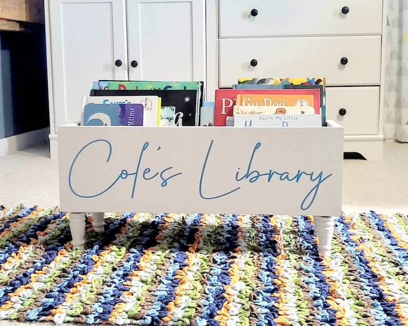 May include: A white wooden bookshelf with the text "Cole's Library" in blue script. The bookshelf is filled with colorful children's books and sits on four white legs. A colorful rug is in the foreground.
