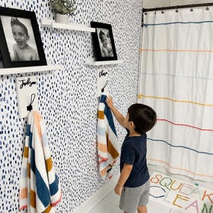 May include: Two white wooden wall-mounted towel hooks with black metal hardware and personalized name plaques, each holding a striped multi-colored towel. Above each hook is a white floating shelf displaying a black framed portrait photograph.