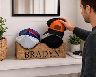 May include: A wooden box with the name "BRADYN" in black lettering holds several baseball caps. The caps are in various colors, including black, orange, blue, and white. A person is reaching for an orange and black cap.