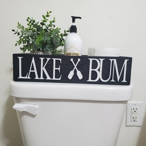 May include: A black wooden sign with white lettering that reads "LAKE X BUM" with crossed oars. The sign is on top of a white toilet, with a small plant, hand lotion, and toilet paper roll.