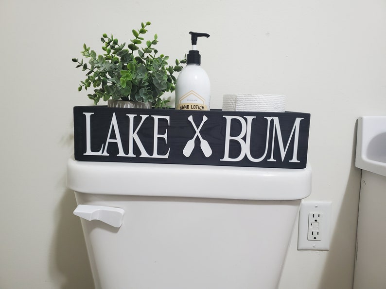 May include: A black wooden sign with white lettering that reads "LAKE X BUM" with crossed oars. The sign is on top of a white toilet, with a small plant, hand lotion, and toilet paper roll.