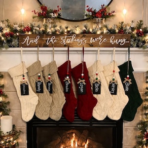 May include: A fireplace mantel decorated for Christmas with seven cable-knit stockings in cream, tan, and red. Each stocking has a name tag. A wooden sign above reads "And the stockings were hung..."