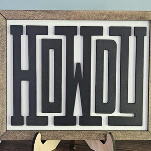 May include: Wooden framed sign with the word "HOWDY" in bold, black letters. The sign has a white background and a brown wooden frame. The sign is displayed on a wooden stand.