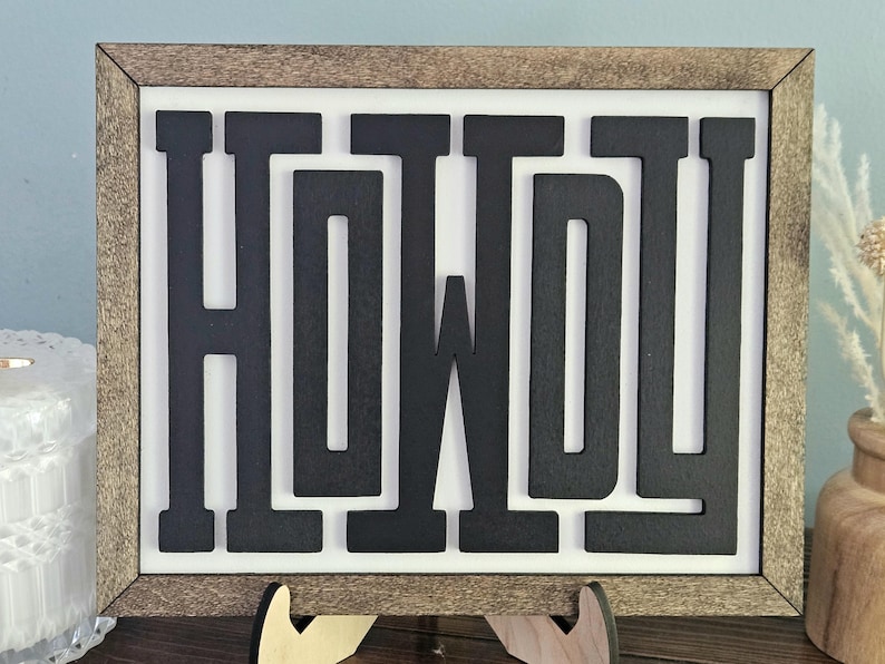 May include: Wooden framed sign with the word "HOWDY" in bold, black letters. The sign has a white background and a brown wooden frame. The sign is displayed on a wooden stand.