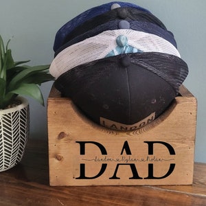 May include: Wooden hat rack with the word "DAD" and the names Landon, Rylan, and Nolan written underneath. The rack is holding a stack of four baseball caps.