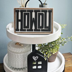 May include: A two-tiered white tray with a black metal handle. The top tier features a wooden sign with the word "HOWDY" in black letters. The bottom tier holds a black house-shaped decoration with a heart design and a glass jar.