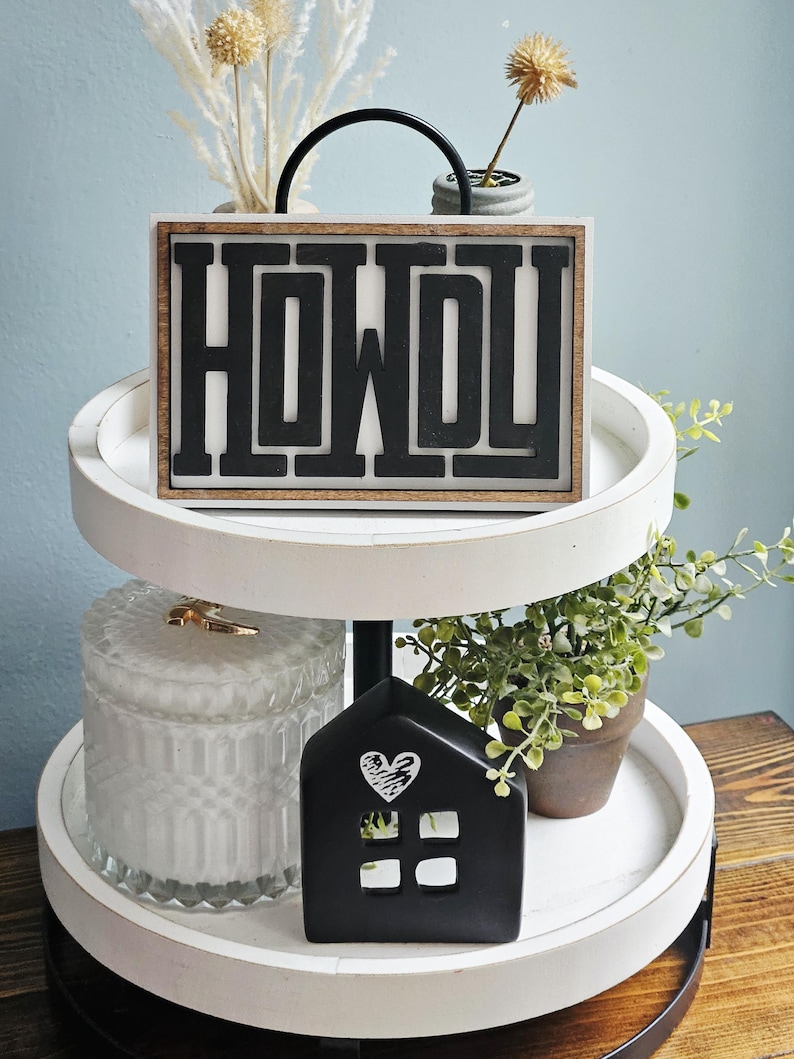 May include: A two-tiered white tray with a black metal handle. The top tier features a wooden sign with the word "HOWDY" in black letters. The bottom tier holds a black house-shaped decoration with a heart design and a glass jar.