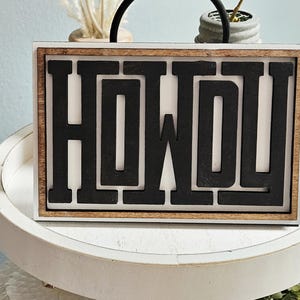 May include: Wooden sign with the word "HOWDY" in black, block letters. The sign has a white background and a brown wooden frame. The sign is sitting on a white, round tray.