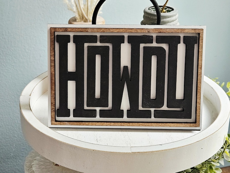 May include: Wooden sign with the word "HOWDY" in black, block letters. The sign has a white background and a brown wooden frame. The sign is sitting on a white, round tray.