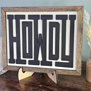 May include: A framed wooden sign with the word "HOWDY" in bold, black letters against a white background. The frame is a light brown color. The sign is propped up on a wooden stand. Decorative items are in the background.