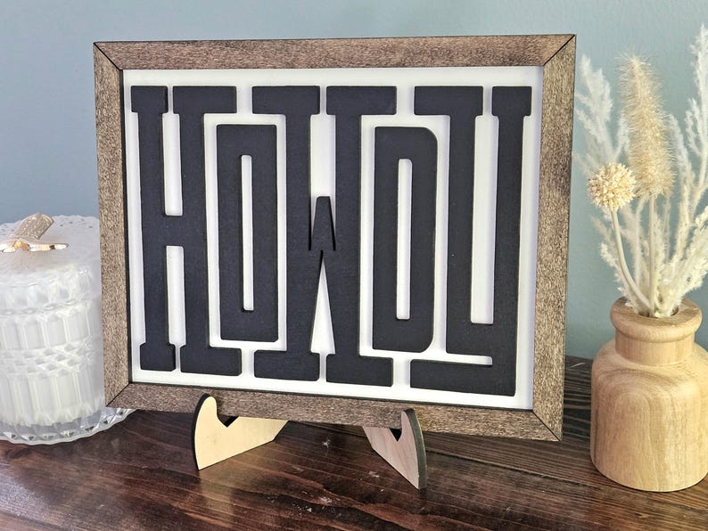 May include: A framed wooden sign with the word "HOWDY" in bold, black letters against a white background. The frame is a light brown color. The sign is propped up on a wooden stand. Decorative items are in the background.