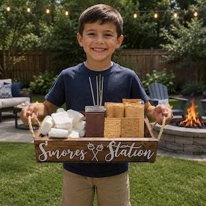 May include: A rectangular dark brown wooden tray with rope handles featuring the white text S'mores Station and a graphic of two crossed roasting sticks. The tray contains a pile of white marshmallows, a container of chocolate, and graham crackers.