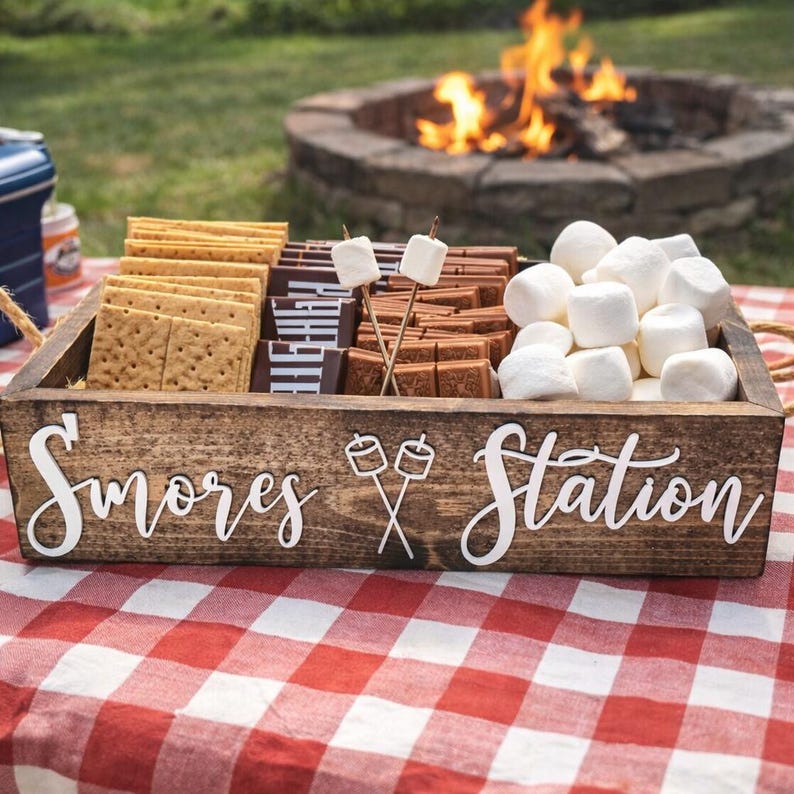 May include: A rectangular wooden box with the white text S'mores Station and a graphic of two crossed marshmallows on sticks. The box contains rows of rectangular graham crackers, dark chocolate bars, and a pile of white marshmallows.