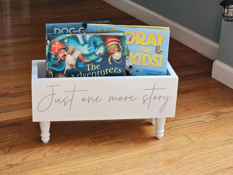 May include: A white wooden book bin with four short, decorative legs. The bin is filled with children's books. The words "Just one more story" are written on the front of the bin in a cursive font.