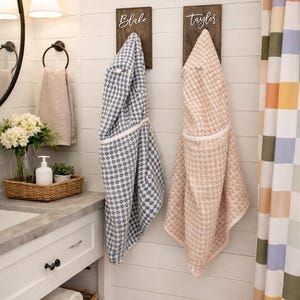 May include: Bathroom scene with two hooded bathrobes hanging on wooden plaques. One is blue and white, the other is pink. The names "Blake" and "Taylor" are written on the plaques. A white sink, mirror, and shower curtain are also visible.