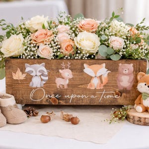 May include: A wooden box with floral arrangements and illustrated woodland creatures. The box reads "Once upon a time." A stuffed fox with a green bow tie sits on a wooden slice. Baby booties and candles are also present.