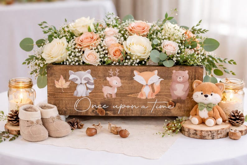 May include: A wooden box with floral arrangements and illustrated woodland creatures. The box reads "Once upon a time." A stuffed fox with a green bow tie sits on a wooden slice. Baby booties and candles are also present.